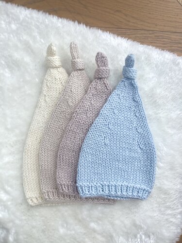 Vier gestrickte Babymützen in Creme, Taupe, Grau-Lila und Hellblau mit Knoten auf weißem Fell.