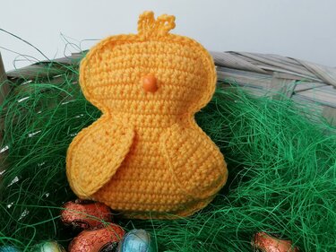 Gehäkeltes gelbes Küken mit orangefarbenem Schnabel in einem Korb mit grünem Gras und bunten Ostereiern.