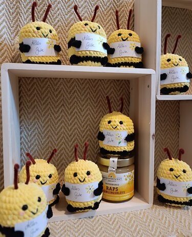 Gehäkelte gelb‑schwarze Bienen‑Amigurumi mit „Alles Gute“-Kärtchen auf hellen Holzregalen.