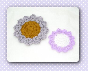 Zwei gehäkelte blumenförmige Deckchen, das größere grau mit senfgelber Mitte, das kleinere lila mit weißer Mitte.