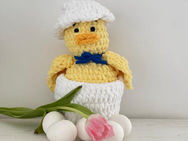 Gehäkeltes gelbes Küken mit weißem Hut und blauer Schleife in weißer Eierschale, mit weißen Eiern und rosa Tulpe im Vordergrund.