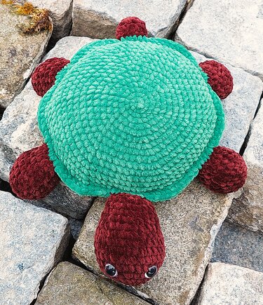 Nahaufnahme einer gehäkelten Amigurumi-Schildkröte mit türkisem Panzer und dunkelrotem Kopf sowie Beinen, liegend auf grauen Felsen.