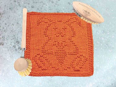 Orangefarbener gestrickter Spüllappen mit Hasenrelief und zwei Holzbürsten auf schaumigem Untergrund.