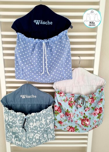 Drei hängende Stoffbeutel in Blau- und Grüntönen mit Punkten, Fisch- und Blumenmuster; zwei mit „Wäsche“-Stickerei, an einem weißen Heizkörper.