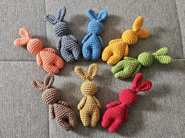 Seven small crocheted amigurumi bunnies in assorted colors arranged in a circle on grey fabric.
