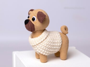 Gehäkelter Mops-Amigurumi in Beige mit braunen Details und cremefarbenem Pullover, seitlich auf weißer Oberfläche