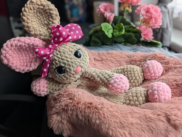 Gehäkelter Amigurumi-Hase in Beige und Rosa mit gepunkteter Schleife, liegend auf rosafarbenem Plüsch