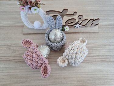 Drei gehäkelte Hasen-Eierwärmer in Grau, Rosa und Creme mit dekorativen Ostereiern auf einem Holztisch