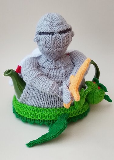 Knitted tea cozy shaped like a grey knight on a green dragon base over a dark green teapot.