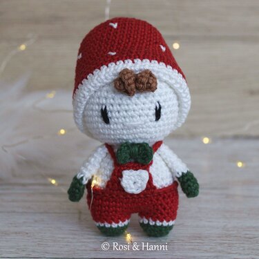 Gehäkeltes Glückspilz‑Amigurumi mit roter Mütze, grüner Fliege und roten Latzhosen vor hellem Hintergrund