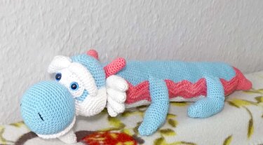 long crocheted dragon amigurumi in pastel blue, white and pink lying on a floral surface