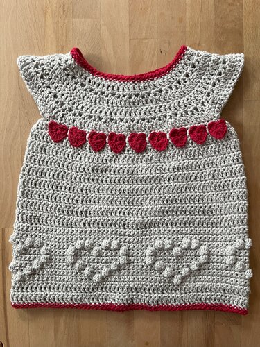 Gehäkeltes hellgraues Babykleid mit roten Herzapplikationen und Noppenmuster auf Holzuntergrund.