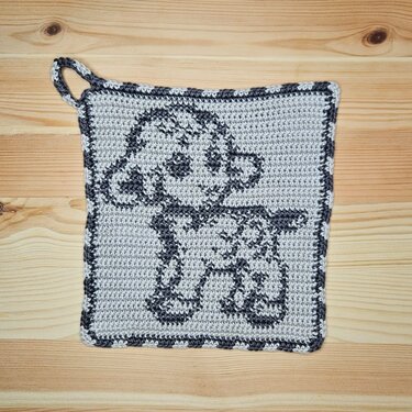 Quadratischer gehäkelter Topflappen mit Lämmchen-Motiv in Hell- und Dunkelgrau und Aufhängeschlaufe auf Holzuntergrund.