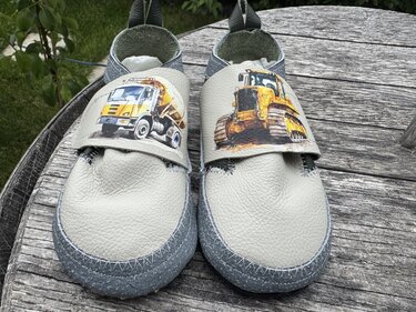 Hellgraue Kinderpuschen aus Leder mit Prints eines Betonmischers und eines Raupenladers auf einer Holzplatte.