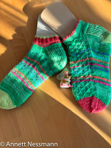Zwei kurzgestrickte grüne Socken mit rosa und blauen Streifen auf Holz-Sockenspannern, liegend auf einem Holztisch, daneben eine kleine Figur
