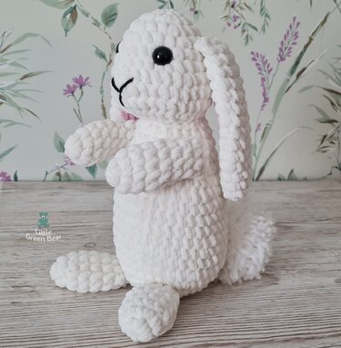 White crocheted amigurumi bunny with black eyes, embroidered nose and floppy ears, sitting on a wooden surface.