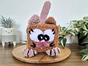 Pastellbrauner gehäkelter Würfel mit rosa Details und großen Augen neben Topfpflanzen