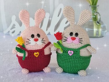 Two crocheted amigurumi rabbits in red and green overalls holding small yarn flowers