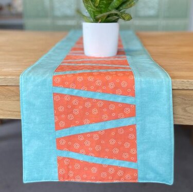 Tischläufer mit türkisfarbenem Rand und orangefarbenem Mittelteil mit diagonalen Patchworkstreifen, auf Holztisch mit Pflanzentopf