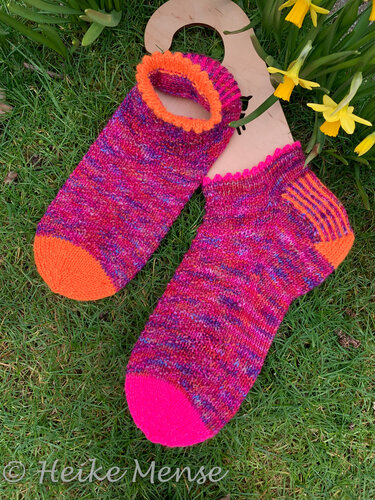 Kurzschaftige, handgestrickte Socken in Pink, Magenta und Orange auf Gras neben Narzissen und einem Sockenbrett