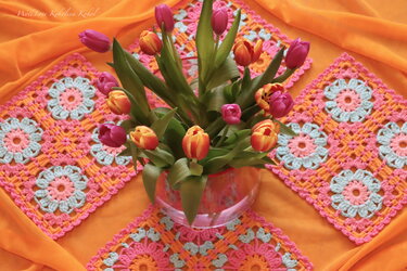Bunter Tulpenstrauß in Glasvase, umgeben von gehäkelten Granny Squares in Pastellfarben auf orangefarbenem Stoff.