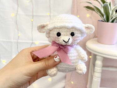 Weißes gehäkeltes Amigurumi-Schaf mit rosa Schleife, in der Hand gehalten
