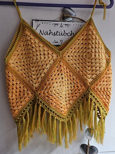 Handgehäkeltes Damen-Top in Orange und Gelb mit grünen Akzenten und Fransen, an einem Kleiderbügel hängend.