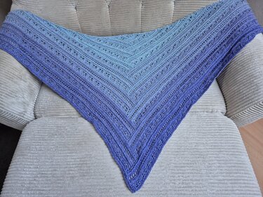 Gehäkeltes Dreieckstuch mit Blau‑Lila‑Farbverlauf und Lochmuster auf hellem, gepolstertem Sessel
