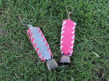 Zwei gehäkelte Schlüsselanhänger, grau mit korallfarbener Naht und pink mit weißer Naht, jeweils mit Metallglocke auf Gras liegend