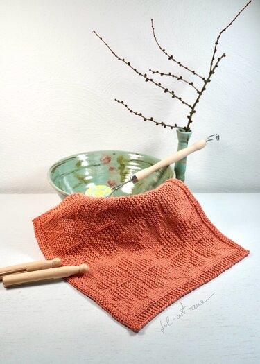 Terrakottafarbenes gestricktes Spültuch mit erhabenem Blattmuster auf hellem Tisch, im Hintergrund Keramikschale und Vase mit Zweig