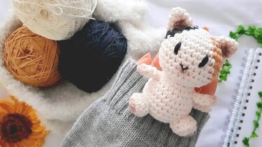 Close-up of a white, orange, and black crocheted amigurumi cat doll next to three balls of yarn, a grey knitted item, and a yellow crochet flower.