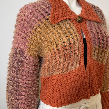 Variegated orange, pink, and brown crocheted women's cardigan with ribbed collar, textured sleeves, and a single round decorative button on a mannequin.