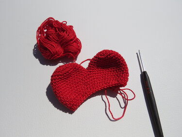 Halb gehäkeltes rotes Herz neben rotem Garn und Häkelnadel auf weißem Hintergrund.