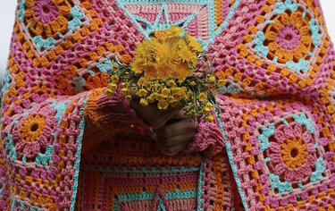 Hände halten einen Strauß gelber Blumen vor einer bunten, gehäkelten Decke mit Granny-Squares in Rosa, Orange und Türkis.