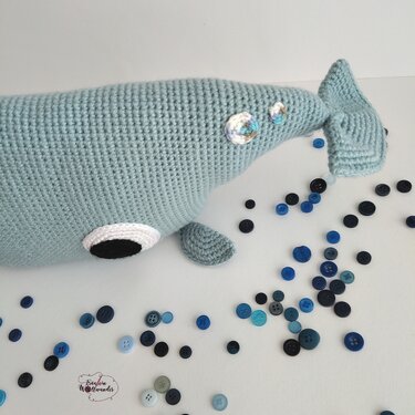 Hellblau gehäkelter Wal Amigurumi liegend, umgeben von blauen Knöpfen