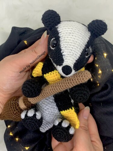 Gehäkelter Dachs Amigurumi mit schwarz-gelbem Schal und kleinem Besen, in Händen gehalten