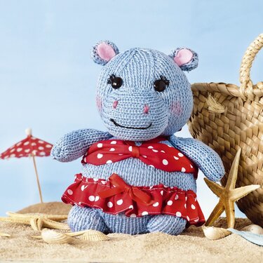 Hellblaues gestricktes Amigurumi-Nilpferd im roten Outfit mit weißen Punkten am Strand mit Muscheln, Seestern und Bastkorb