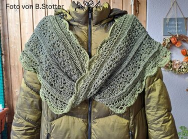 Grüner gehäkelter Loopschal mit offenem Lochmuster und gewellter Kante, lässig über einer khakigrünen Steppjacke drapiert.