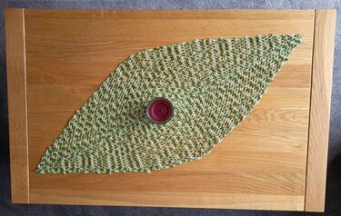 Rautenförmiger, grün-weiß gehäkelter Tischläufer mit Lochmuster auf einem hellen Holztisch, mittig eine dunkelrote Kerze in einem Glas.