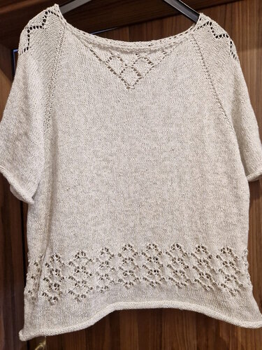 Helles gestricktes Damen-Kurzarmshirt mit Lochmuster am Saum und Rautendetail am Ausschnitt, auf einem Bügel hängend.