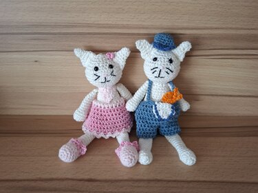 Zwei gehäkelte Katzen-Amigurumi nebeneinander, links rosa Kleid, rechts blaue Latzhose mit Hut und orangefarbener Karotte in der Tasche