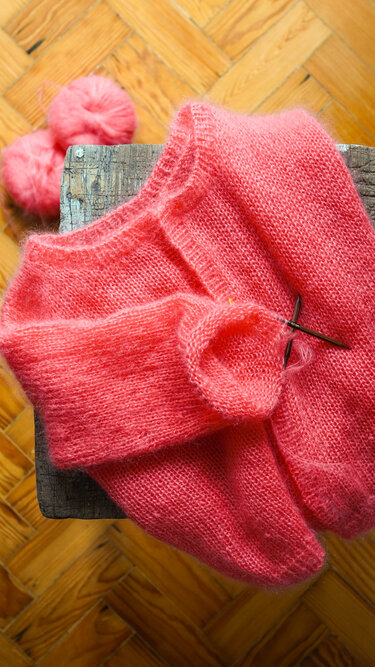 Unfertige korallenrote Strickjacke mit Stricknadeln und Wollknäueln auf Holzhocker