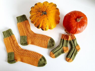Zwei Paare gestrickter Socken in Orange und Oliv mit Ajourmuster neben zwei Kürbissen