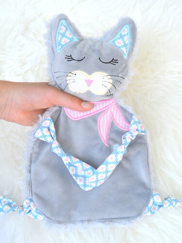Graues Katzen-Schmusetuch mit gesticktem Gesicht, rosa Halstuch und gemusterten Knoten auf hellem Hintergrund.