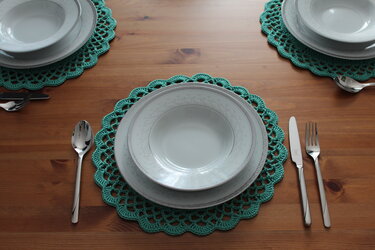 Drei türkisfarbene gehäkelte Tischsets auf einem Holztisch, mit weißen Tellern und Besteck.