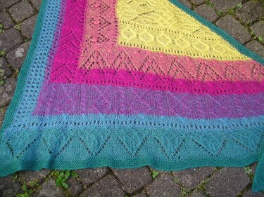 Gestricktes Dreieckstuch mit Farbverlauf in Gelb, Pink, Lila und Türkis mit Loch- und Zopfmustern auf Pflastersteinen.