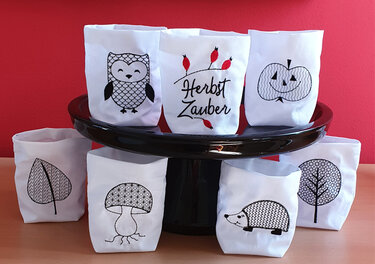 Sieben weiße bestickte ITH-Teelichthüllen mit Eule, Schriftzug 'Herbst Zauber', Kürbis, Blatt, Pilz, Igel und Baum vor einer roten Wand