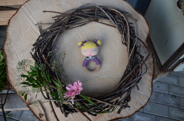 Gehäkelte Baby-Rassel mit gelben Zöpfchen und lila Oberteil auf Holzring, platziert auf einer Baumscheibe mit Zweigkranz und rosa Blüte