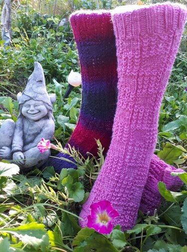 Zwei gestrickte Kniestrümpfe, rosa und mehrfarbig, im Gras neben einem lächelnden Gartenzwerg
