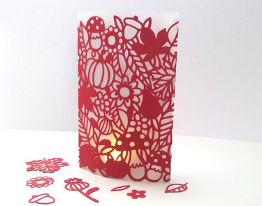 Rote, fein geschnittene Papierlaterne mit Pilz-, Blumen- und Blattmotiven, innen beleuchtet; ausgeschnittene Dekoteile liegen auf weißem Untergrund